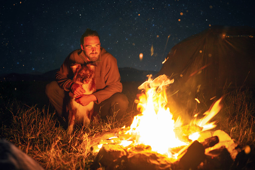 A bearded man in his 20s is sitting behind a bonfire with his dog in front of him. Star filled sky in the background.
