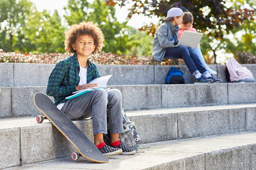 Portrait of African boy with curly hair sitting on stairs with skateboard and smiling at camera while reading a book with children in the background