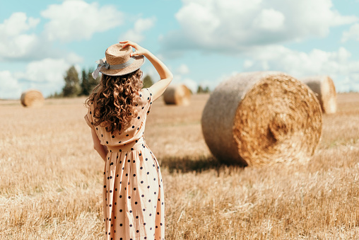 Woman in beige polka dot dress with curly hair, straw hat standing on harvested field with straw bales. Agriculture background with copy space