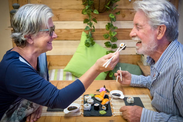 alegre pareja de ancianos, un hombre y una mujer con canas, disfrutando del sushi japonés. al aire libre sobre una mesa de madera - sushi restaurant fish japanese culture fotografías e imágenes de stock