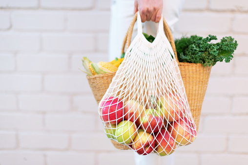 Girl holding mesh shopping bag full of apples and straw bag with organic vegetables, brick background. Zero waste, plastic free concept. Sustainable lifestyle. Copy space