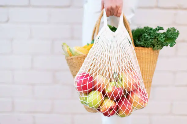 Photo of Girl holding mesh shopping bag full of apples and straw bag with organic vegetables, brick background. Zero waste, plastic free concept. Sustainable lifestyle. Copy space