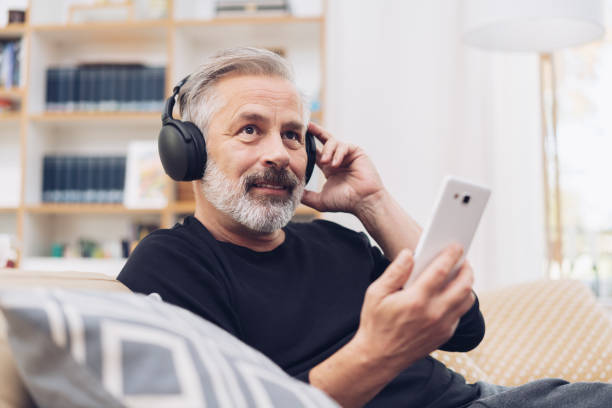 Middle-aged man listening to music online at home Portrait of a middle-aged man using his mobile phone for listening to music online through modern headphones at home podcast mobile stock pictures, royalty-free photos & images