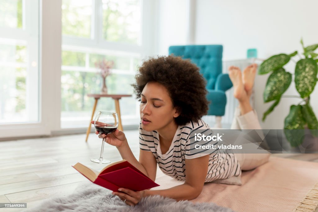 Woman reading a book and drinking wine Portrait of a beautiful young mixed race woman lying on the living room floor, drinking wine, reading a book and relaxing at home Book Stock Photo