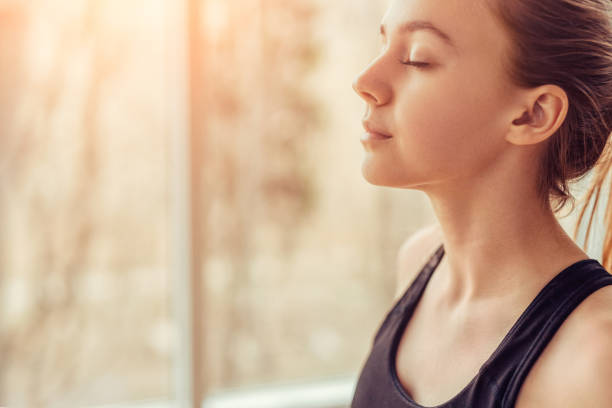 Young woman doing breathing exercise Side view of young female with closed eyes breathing deeply while doing respiration exercise during yoga session in gym deep stock pictures, royalty-free photos & images