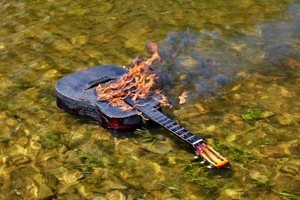 They set the guitar on fire and put it in water. An ignited guitar floats on riverbank, fire burns on its surface. From the flame comes black smoke. The strings are torn. A musical instrument drifts along a river, it is charred and destroyed by fire Aflame stock pictures, royalty-free photos & images