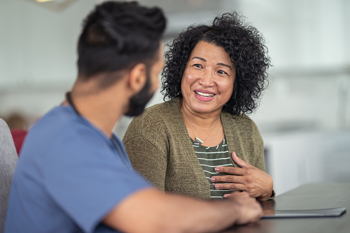 A woman of Asian descent is at a medical consultation. She is sitting at a table next to her doctor. The doctor is a mixed-race man of Asian and Indian descent. The two individuals are having a conversation. The patient is smiling and talking.