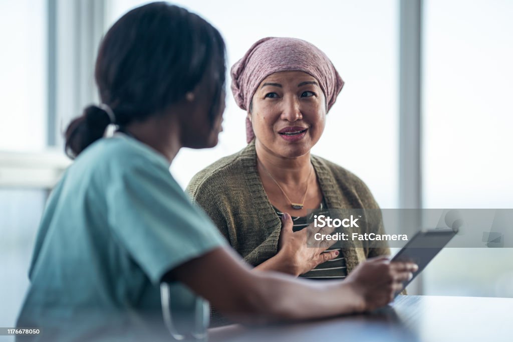 Asian woman with cancer meeting with female physician An Asian woman with cancer is consulting her doctor. The two women are seated at a table together. The patient is wearing a bandana to hide her hair loss. The medical professional is showing the patient test results on a digital tablet. They are discussing a treatment plan. Cancer - Illness Stock Photo