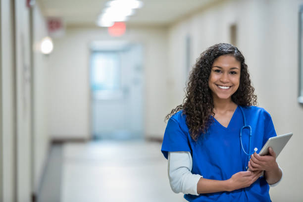 healthcare worker standing in hospital hallway holding a digital tablet - female nurse imagens e fotografias de stock