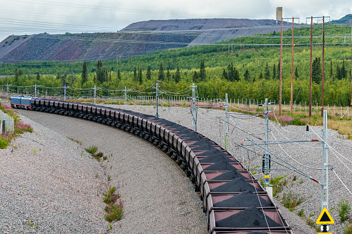 Looking down on a loaded electric iron ore train passing mine tailings on a gloomy day. Train is going away from the camera showing neat piles of loaded iron ore in the railroad cars. All logos and ID removed.