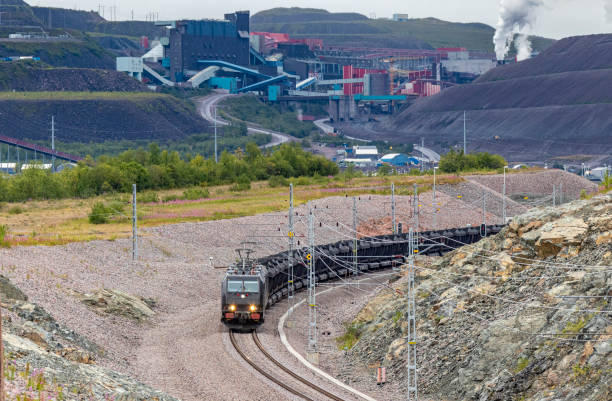 Loaded electric iron ore train passing mine operations Looking down on a loaded electric iron ore train passing mine operations in the background. The all-black train is curving around a rocky embankment with multi-colored mine buildings behind. All logos and ID removed. norrbotten province stock pictures, royalty-free photos & images