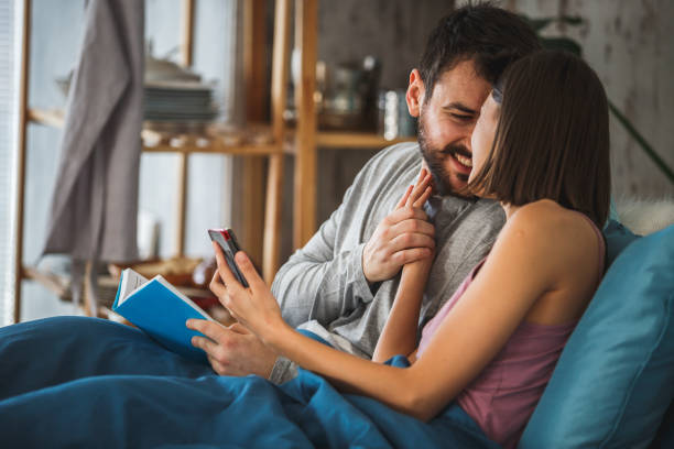 istock Happy young married couple having a cuddle in bed while reading a book and using smart phone 1176710232