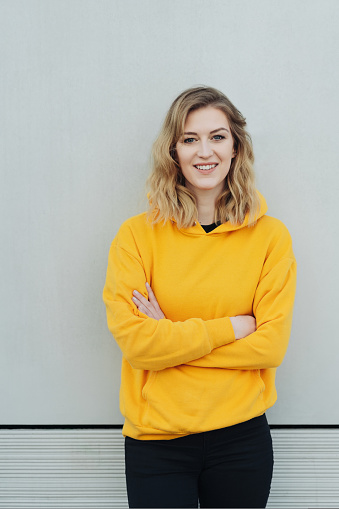 Relaxed young woman in a colorful yellow top standing in front of a white wall with folded arms looking at camera with a friendly smile