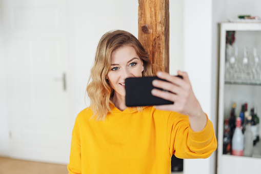Smiling young woman taking a selfie at home to share on social media with focus past the phone to her face