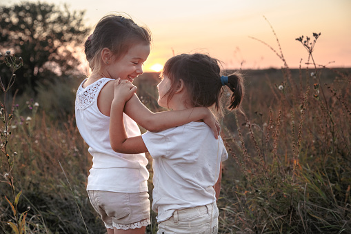 Two little sisters hugging in a field at sunset . Dressed in white. The concept of family values and friendship .