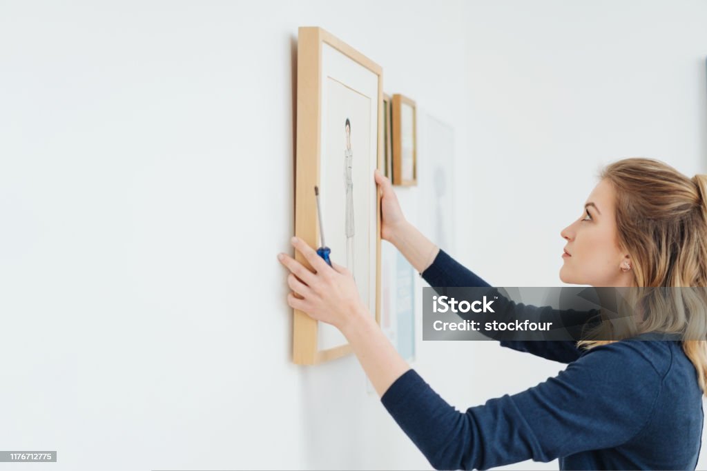Young woman hanging a picture on a wall Young attractive blond woman hanging a picture in a plain wooden frame on a wall with a look of concentration and copy space Hanging Stock Photo