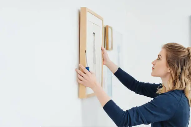 Young attractive blond woman hanging a picture in a plain wooden frame on a wall with a look of concentration and copy space