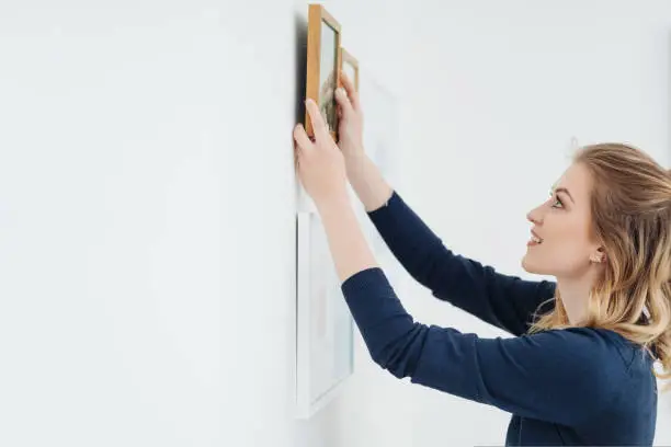 Young woman with a smile decorating her home hanging pictures on a white interior wall with copy space