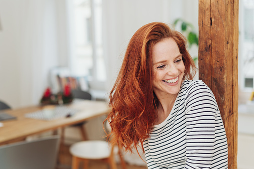 Laughing carefree young woman indoors at home leaning against an old wooden pillar looking to the side smiling