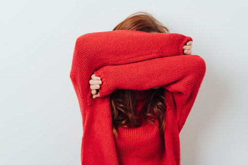 Redhead woman in a colorful red outfit standing hiding her face with her arms over white