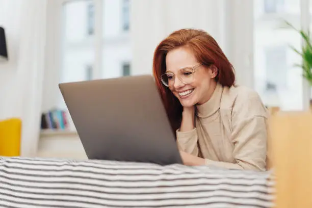 Young woman working on her laptop with a beaming smile as she relaxes on a day bed at home in a low angle view