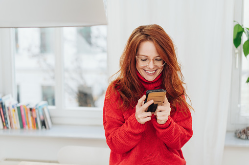 Happy young redhead woman reading on her mobile phone with a pleased smile indoors in her apartment with copy space