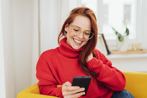 Happy beautiful red-haired girl in glasses and red sweater looking at smartphone in her hand, sitting at home in yellow armchair and smiling