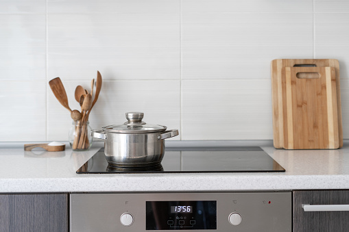 Black ceramic induction stove with timer on control panel and saucepan on top. Contemporary home with modern interior, built in kitchen appliance and white tile on wall with copy space