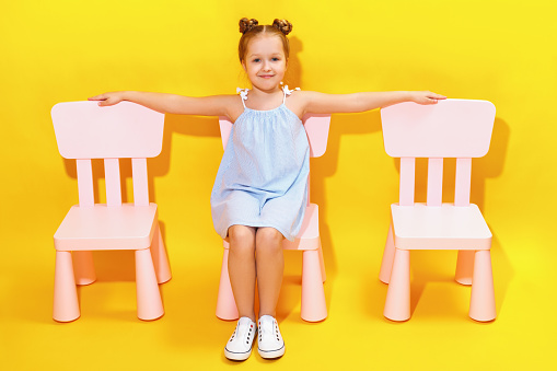 Happy little girl with outstretched arms sits on a chair against the background of a yellow wall. There are empty chairs on the sides of the child. Childhood concept.
