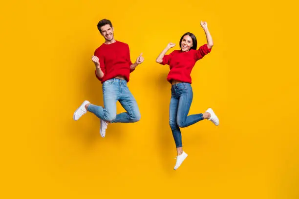 Photo of Full length photo of cheerful woman and man jump raise fists scream yes celebrate victory wear red sweater denim jeans isolated over yellow background