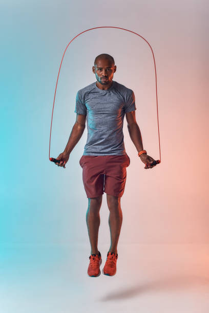 rester en forme. pleine longueur de jeune homme africain dans la corde de saut de vêtements de sport tout en exerçant dans le studio sur le fond coloré - staying fit photos et images de collection