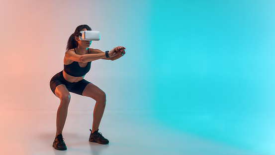 Full length of young sporty woman in sports clothing doing squat while wearing virtual reality glasses. Studio shot. Future technology