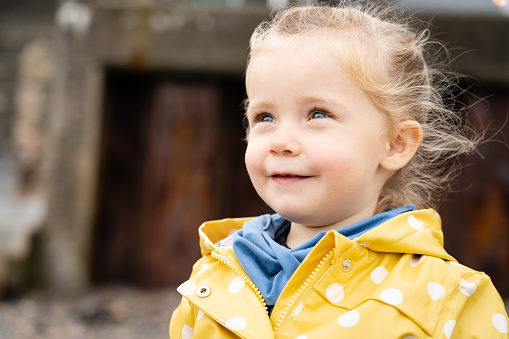 A 2 year old girl with a yellow raincoat smiles outdoors