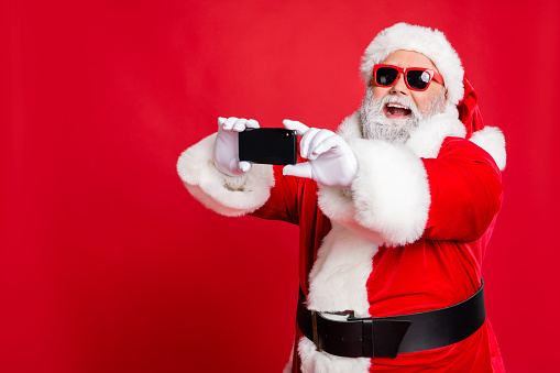 Portrait of excited santa claus in eyewear making selfie with cell phone, device on christmas vacation smiling wearing belt isolated over red background