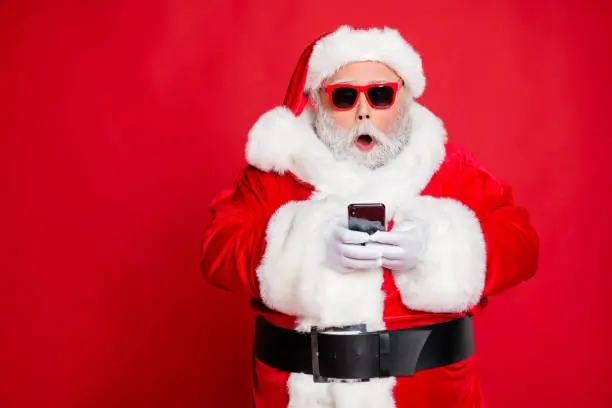 Portrait of impressed christmas father in hat using cell phone device, screaming omg wearing read about festive events eyeglasses eyewear belt isolated over red background