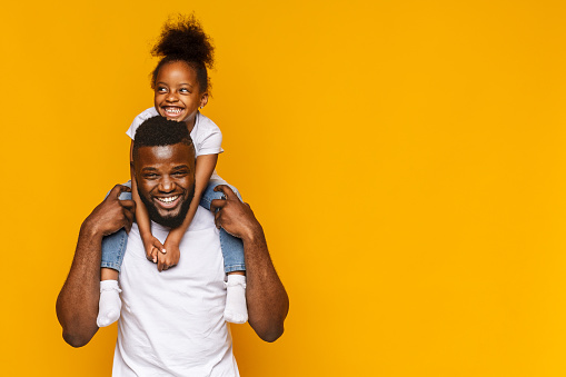 Happy african american father and daughter having fun, riding on shoulders, orange background with copy space