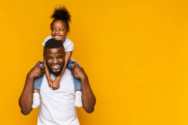 Happy african american father and daughter having fun Happy african american father and daughter having fun, riding on shoulders, orange background with copy space african american children stock pictures, royalty-free photos & images