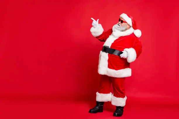 Full length profile side photo of charming santa claus in eyewear eyeglasses showing promo wearing bright costume belt isolated over, red background