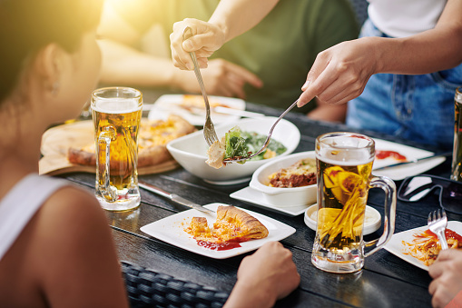 Close-up of young people sitting at the table and have dinner they eating pizza and salad and drinking beer