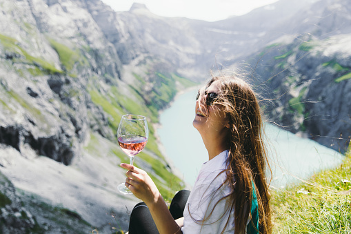 Young smiling woman with long hair in sunglasses resting after a long hiking trip in the mountains, drinking a red wine from the glass and enjoying the view of a lake and mountains in Switzerland