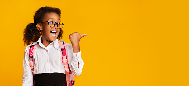 School Offer. Excited Black First-Grade Schoolgirl Pointing Thumbs At Copy Space On Yellow Background In Studio. Panorama