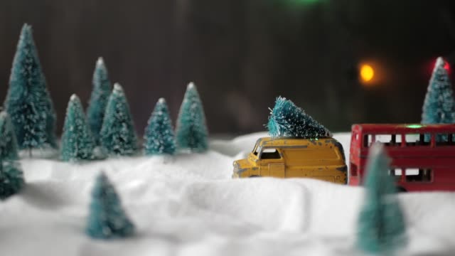 A small toy yellow toy car with a Christmas tree on the roof slowly rides through a miniature toy forest with snowdrifts and Christmas trees.