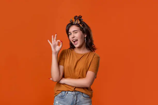 Smiling brunette woman in an orange t-shirt and beautiful headband showing OK-sign with fingers and winking at the camera standing isolated over orange background. Concept of positive evaluation.