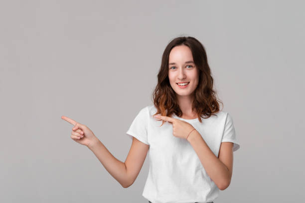 istock Pretty brunette woman in a white tee pointing at the empty space in a left side of a frame standing isolated over grey background. 1176780632
