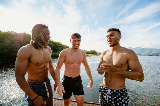 Three young multi ethnic guys in swimwear talking on a jetty in Derwent Water in Cumbria