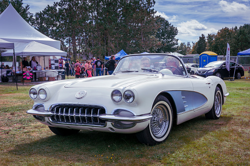Waterville, Nova Scotia, Canada - September 14, 2019 : Driver & passenger in a 1959 Chevy Corvette leave Rick Rood's Car Show in Annapolis Valley. In the background walk about at the classic car show.