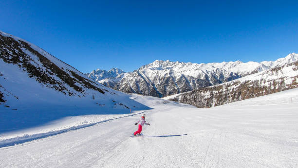A snowboarder going down the slope in Heiligenblut, Austria. Perfectly groomed slopes. High mountains surrounding the man wearing yellow trousers and blue jacket. Girl wears helm for the protection.