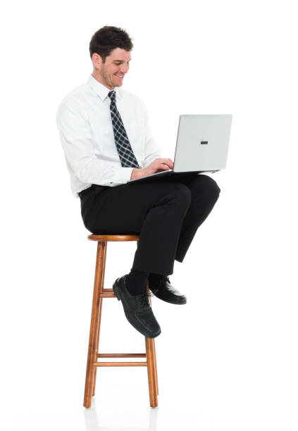 Full length / one man only / one person / side view / profile view of 30-39 years old handsome people caucasian male / mid adult men / mid adult businessman / business person / manager sitting / resting in front of white background wearing button down shirt / shirt / necktie / pants who is smiling / happy / cheerful who is working and using laptop / computer / sitting on stool longitud completa / un hombre solamente / una persona / vista lateral / vista de perfil de 30-39 años de edad personas guapos hombres caucásicos / hombres adultos medios / hombre de negocios de adultos medios / persona de negocios / gerente sentado / des