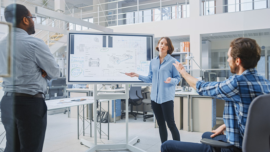 In Industrial Design Facility Team of Engineers and Technicians have a Meeting, Female Specialist Leads Briefing, Talks and Draws on Digital Interactive Whiteboard with Car Prototype Concepts