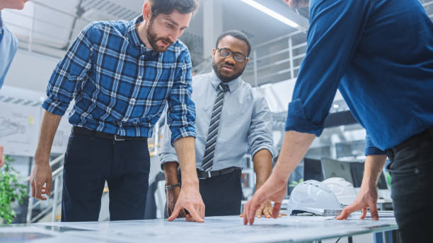 istock In the Industrial Engineering Facility: Diverse Group of Engineers and Technicians on a Meeting Gather Around Table Unravel Sheets of Engine Design Technical Drafts, Have Discussion, Analyse Drawings 1176842999